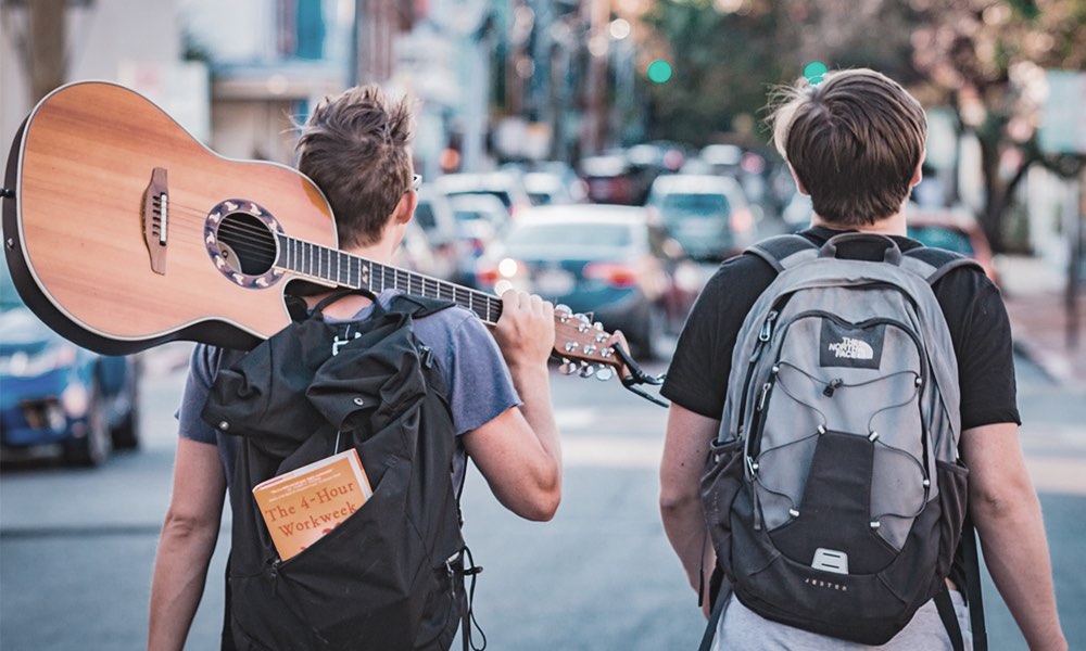 young men carrying a guitar, walking beside a busy street